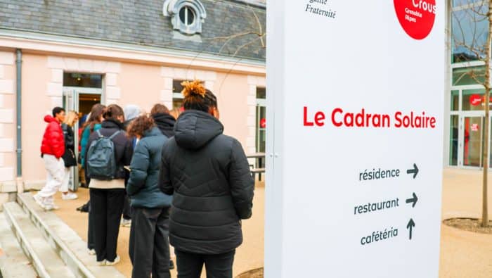 étudiantes et étudiants faisant la queue devant une cafétéria Crous. En premier plan un panneau indiquant Cadran Solaire avec les directions vers cafétéria, restaurant et résidence.