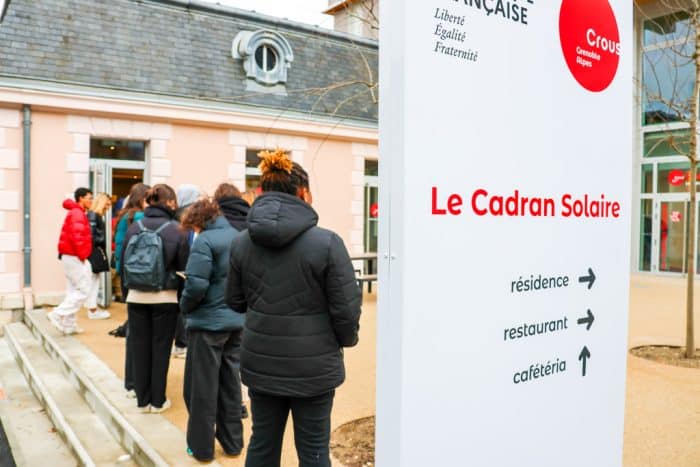 étudiantes et étudiants faisant la queue devant une cafétéria Crous. En premier plan un panneau indiquant Cadran Solaire avec les directions vers cafétéria, restaurant et résidence.